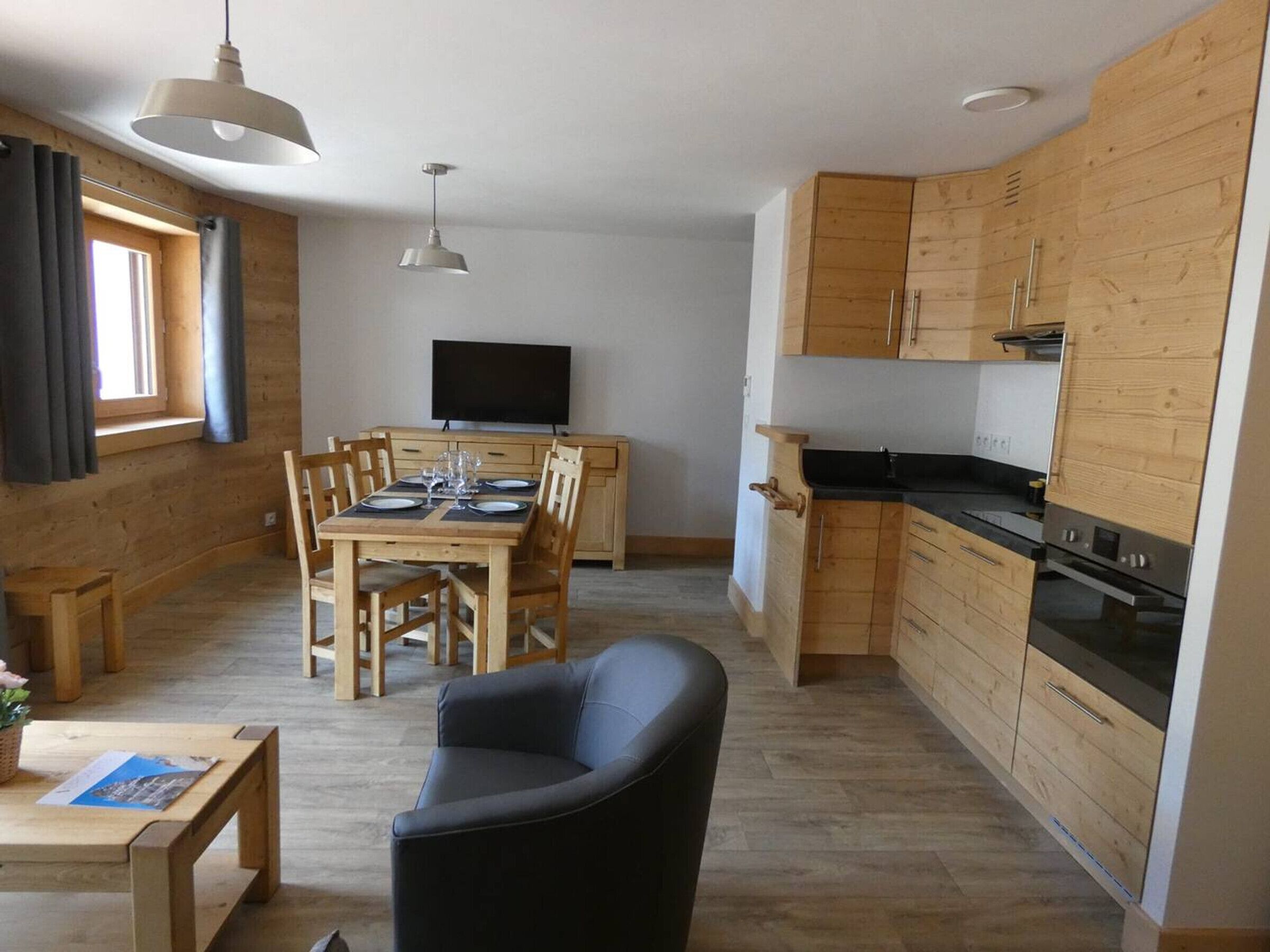 A modern kitchen with wooden cabinets, a dining area with a table and chairs, a black sofa, and a TV mounted on the wall.