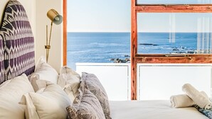 Un dormitorio con un ventanal grande con vista al mar, una cama con almohadillas estampadas y una lámpara de noche.
