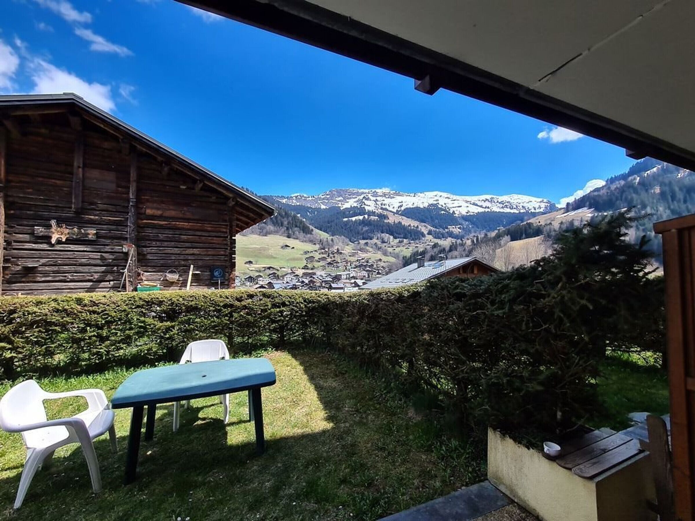 Una cabaña de madera con mesa y sillas en el exterior, con vistas a un pueblo de montaña y picos nevados.