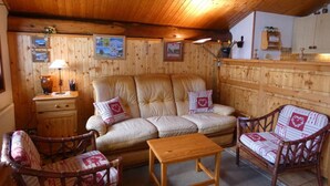 Ein gemütliches Zimmer mit einer Couch, zwei Sesseln, einem kleinen Tisch, Holzwänden, einem Schrank und einer Lampe.