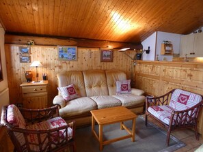 Ein gemütliches Zimmer mit einer Couch, zwei Sesseln, einem kleinen Tisch, Holzwänden, einem Schrank und einer Lampe.