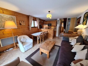 A cozy cabin interior with a wooden dining table, a sofa, a chair, a lamp, and a kitchen area.