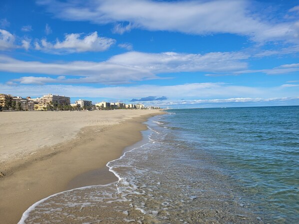 Ein Strand mit klarem Wasser, einem Sandufer und Gebäuden im Hintergrund.
