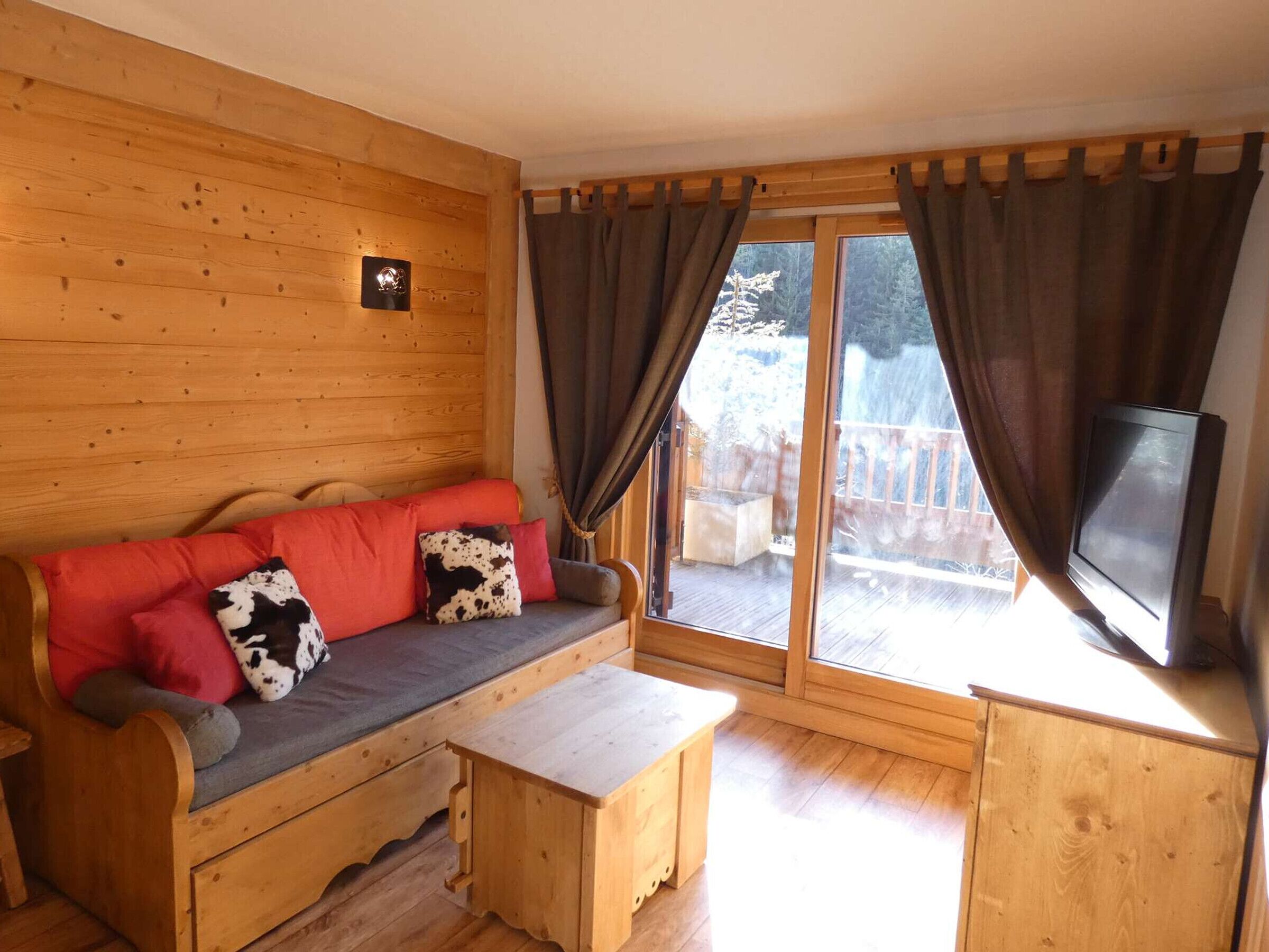 Interior de uma cabana de madeira com sofá, televisão e uma porta de correr para um deck externo.