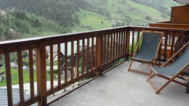 Een balkon met houten hekwerk, twee opklapbare stoelen en uitzicht op een vallei met huizen en een rivier.