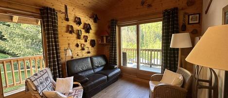 L’intérieur d’un chalet en bois est aménagé avec un canapé en cuir noir, deux fauteuils en osier, un plancher en bois et un porte-chaussures fixé au mur.