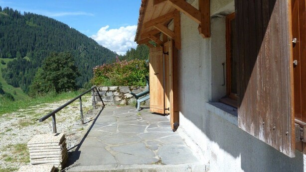 Eine Steintreppe führt zu einem Holzhaus mit einer Steinmauer und Blick auf einen bewaldeten Berg.