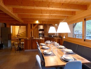 A wooden interior with a long dining table set for six, a kitchen area, and a view through large windows. - Spacious chalet with parking - 13 guests (Arêches-Beaufort)