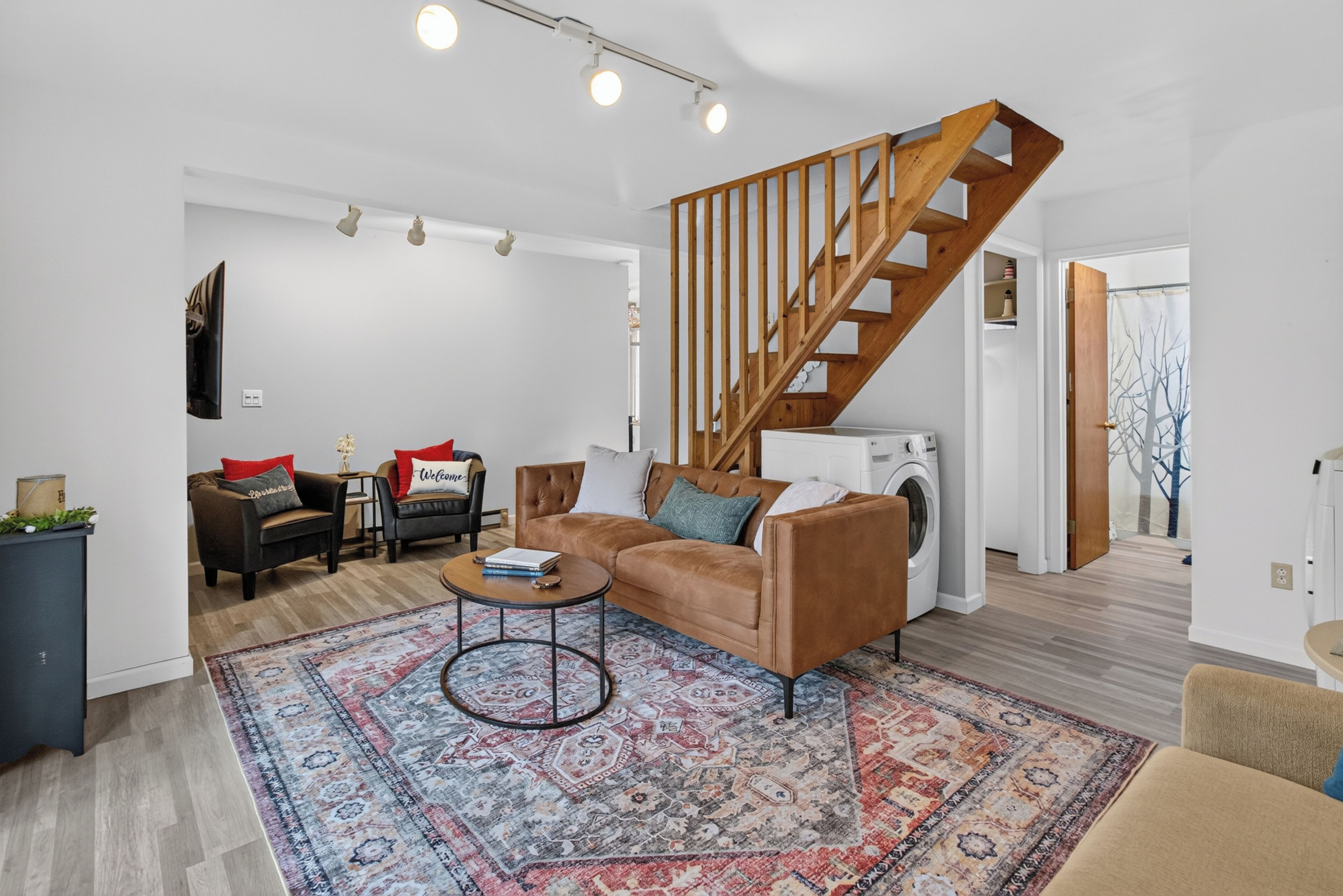 Ein Wohnzimmer mit braunem Ledersofa, einem gemusterten Teppich, einer Holztreppe und einer Waschmaschine im Hintergrund.