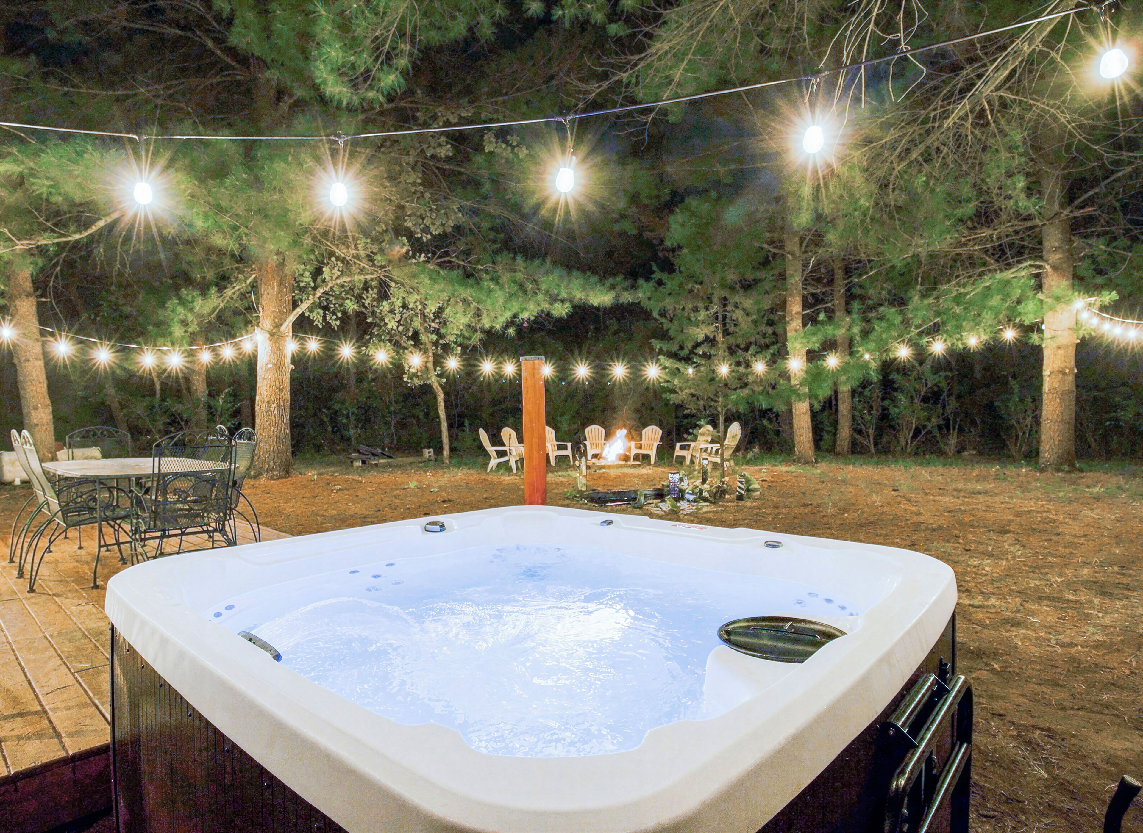 A hot tub surrounded by outdoor furniture and string lights in a wooded area.
