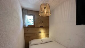 A small, cozy room with a single bed, a wooden ceiling, a window with curtains, and a hanging light fixture. - Cozy apartment in village center - 4 guests (Arêches-Beaufort)