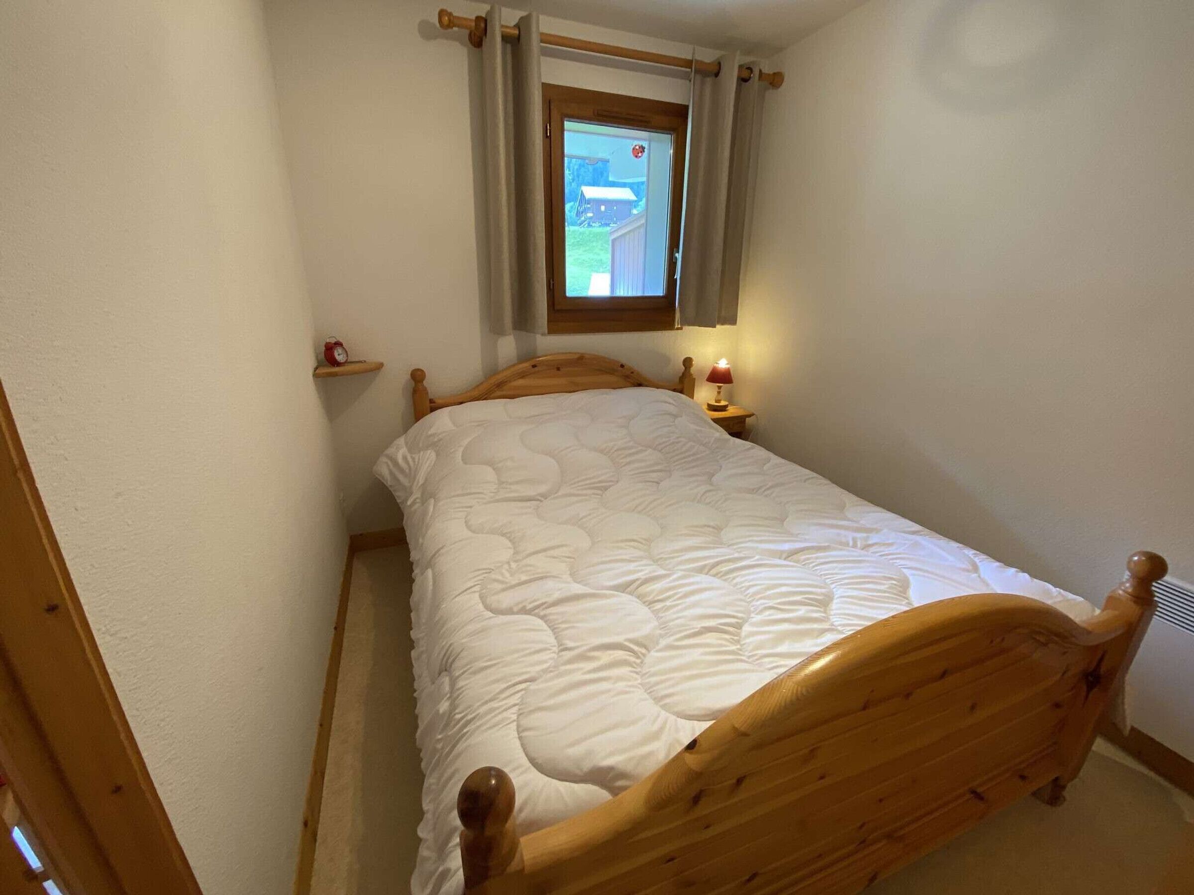 Una stanza piccola con un letto singolo, una finestra con tende, un comodino con una lampada e una mensola con una sveglia.