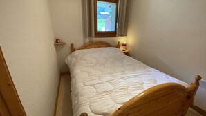 Una habitación pequeña con una cama individual, una ventana con cortinas, una mesita de noche con lámpara y un estante con un reloj.