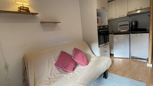Un dormitorio con cama, lámpara de noche, espacio de cocina con electrodomésticos y un estante pequeño.