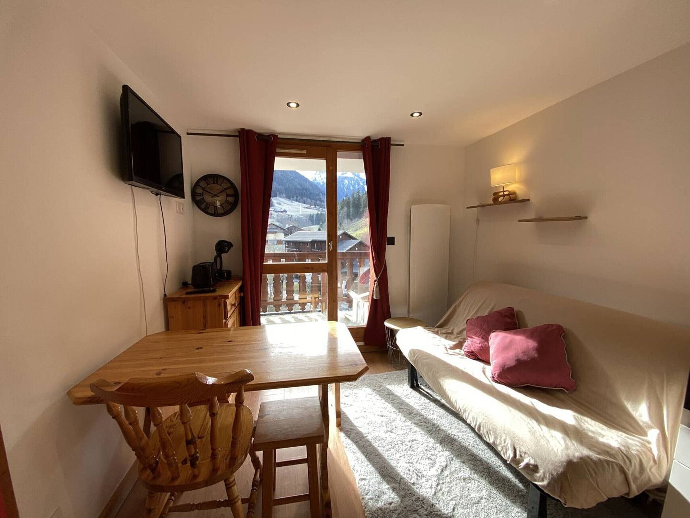 Een kamer met een houten tafel, een bank, een aan de muur bevestigde televisie en uitzicht op bergen door een raam.