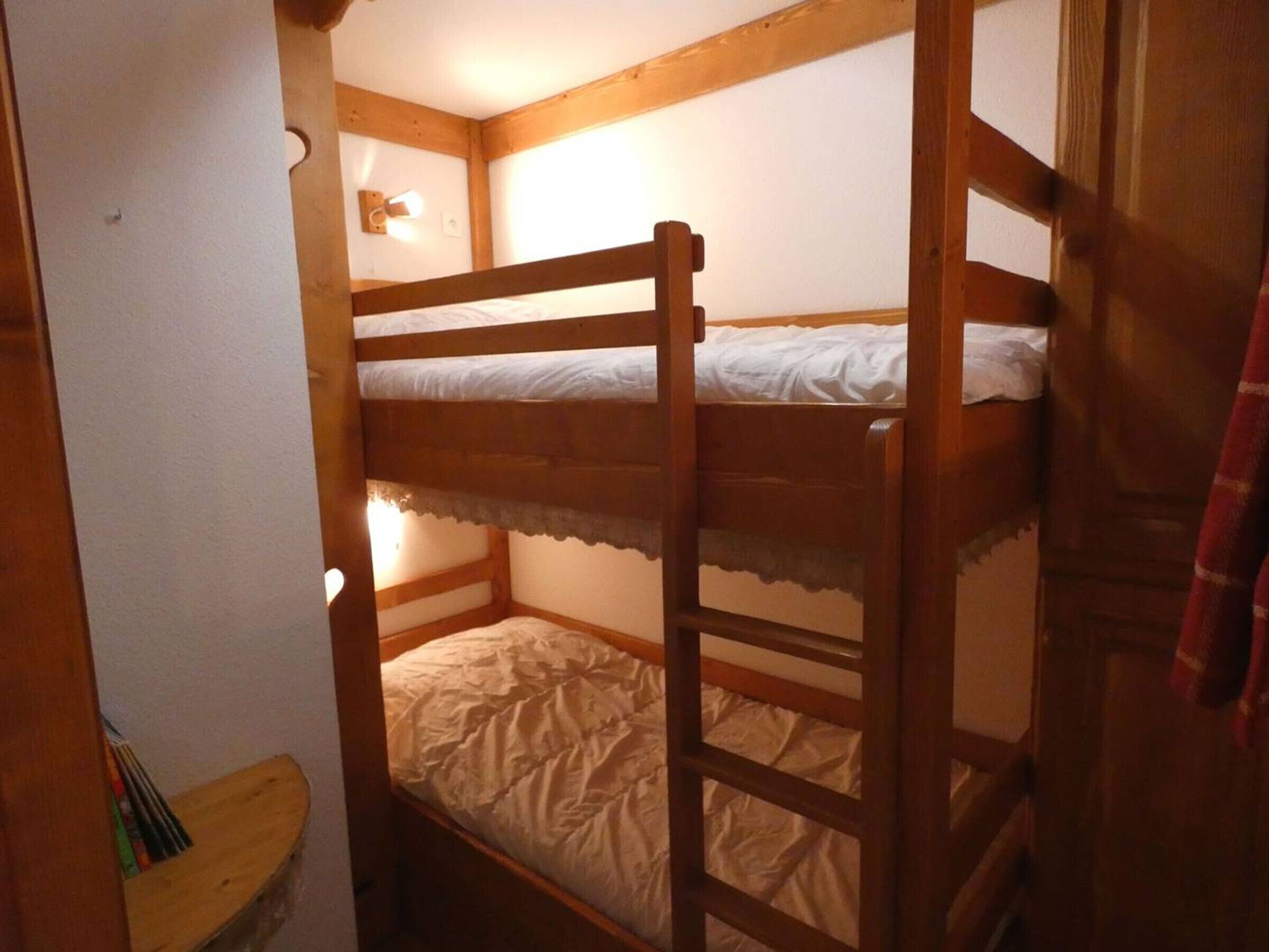 Une chambre avec un lit superposé, des poutres en bois, une petite table avec des livres et un luminaire fixé au mur.