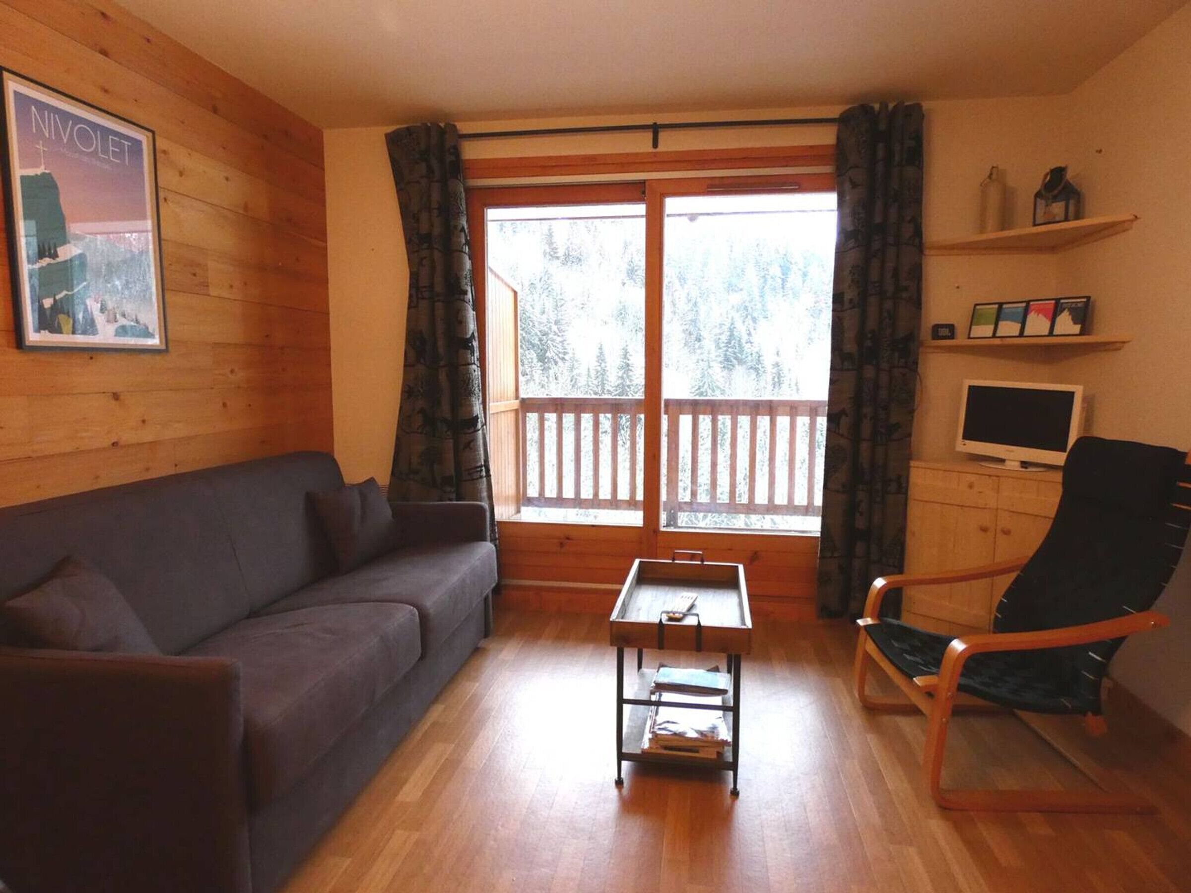 A cozy room with a wooden floor, a sofa, a table, a chair, a TV, and a window with curtains.