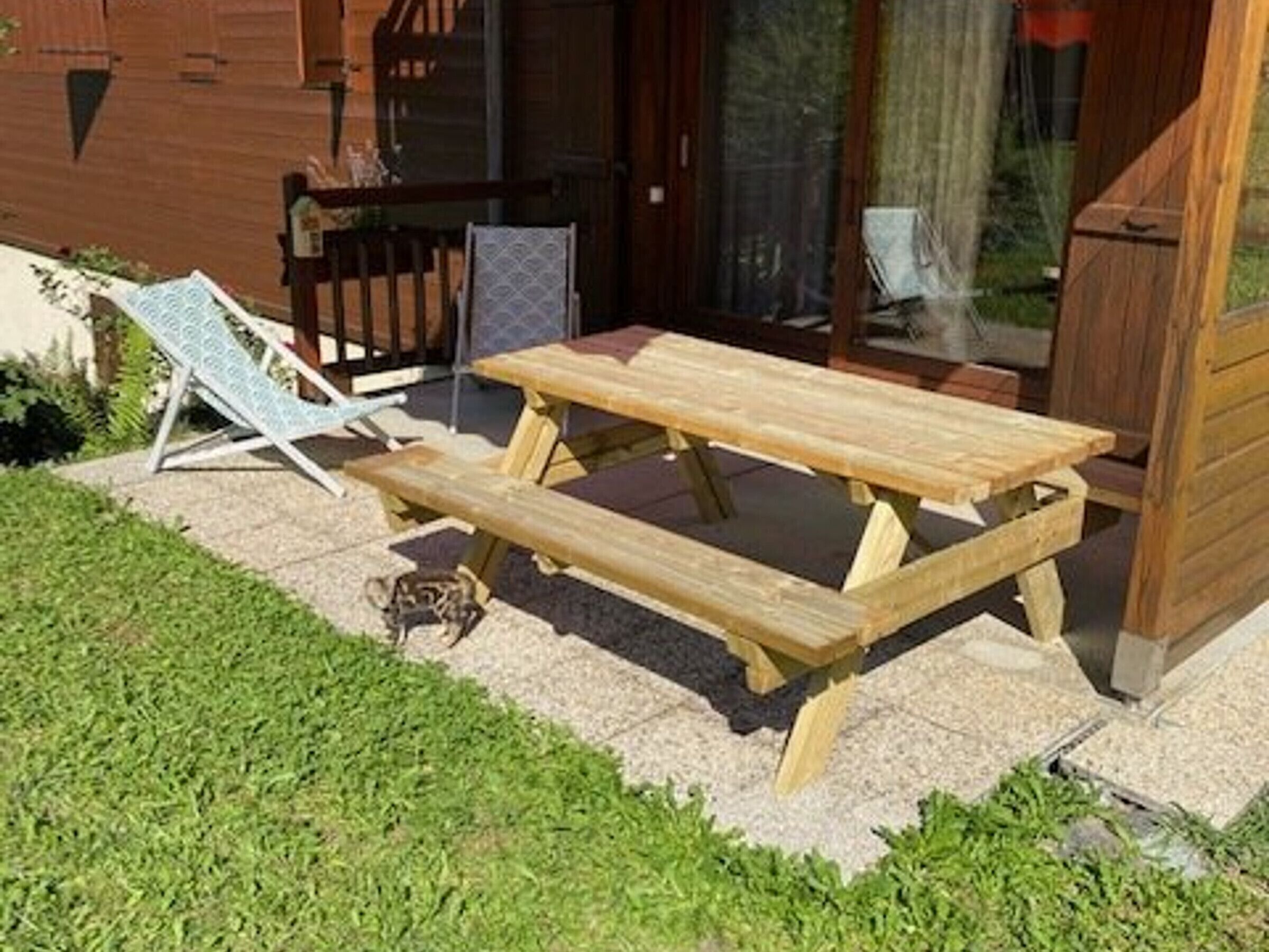 A wooden picnic table with a bench, a lounge chair, and a wooden fence.