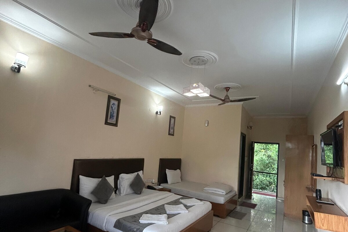 A hotel room with two beds, a sofa, a coffee table, ceiling fans, a TV, and a view of greenery outside.