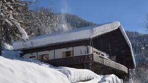 Et snedækket træchalet med balkon, foran et bjerglandskab.