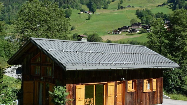 Une cabane en bois avec un toit en pente, de grandes fenêtres et une vue sur une vallée verdoyante et des collines boisées.