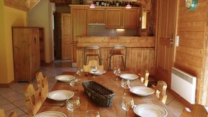 Une table de salle à manger en bois, mise avec des assiettes, des verres et un panier, dans une pièce aux murs en bois, avec une cuisine en arrière-plan.