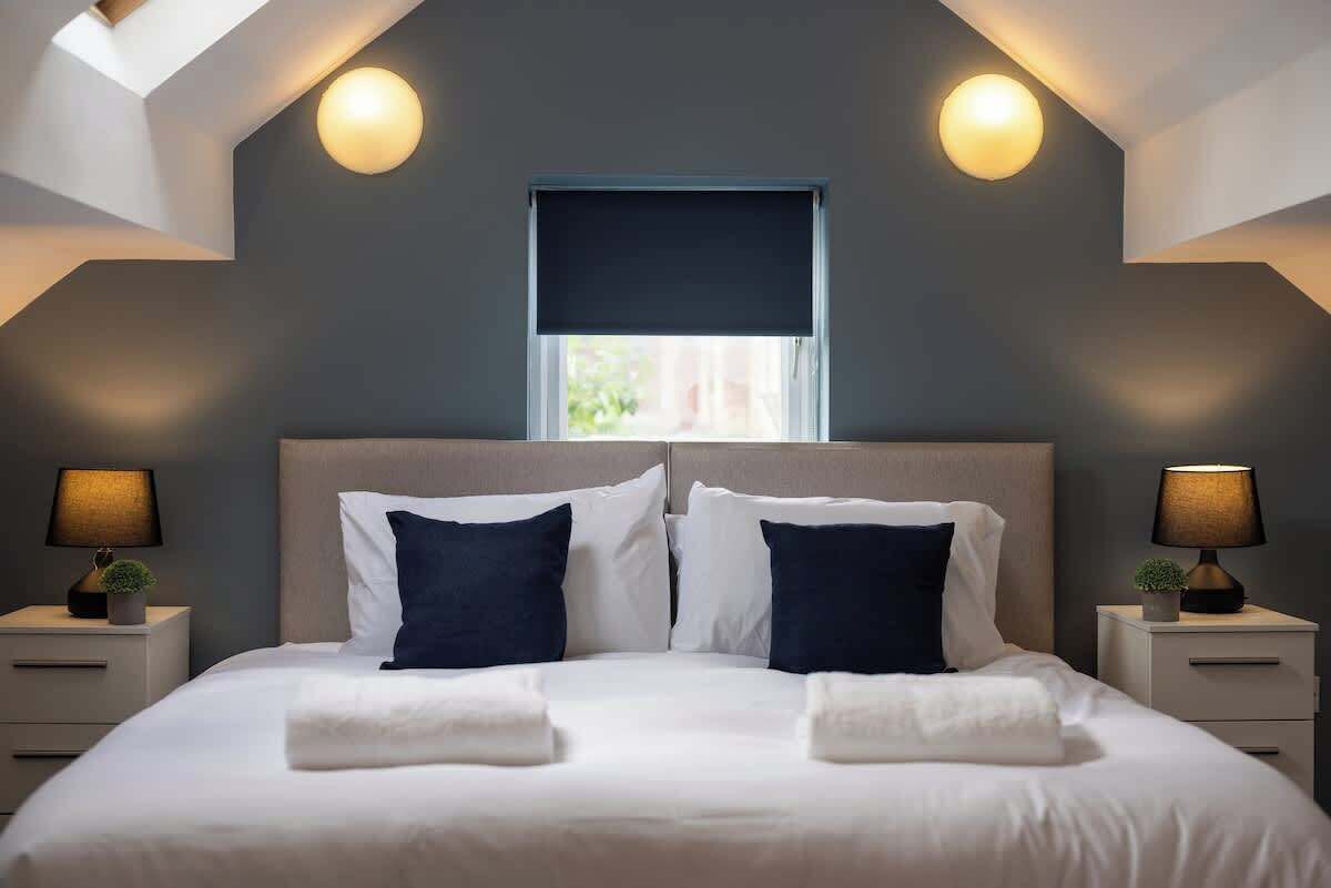 Un dormitorio con cama, dos mesitas de noche, lámparas, una ventana con cortina y un tragaluz.