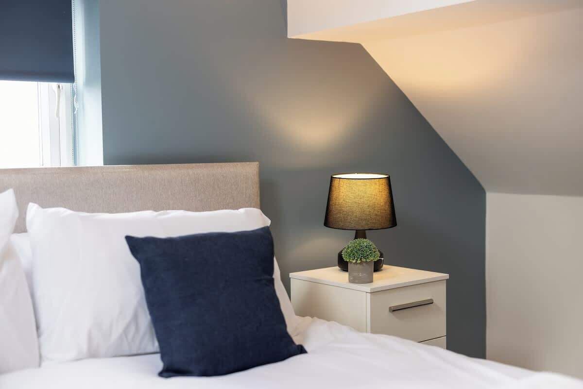 A neatly made bed with white and dark blue pillows, a bedside lamp, and a small plant on a white nightstand.