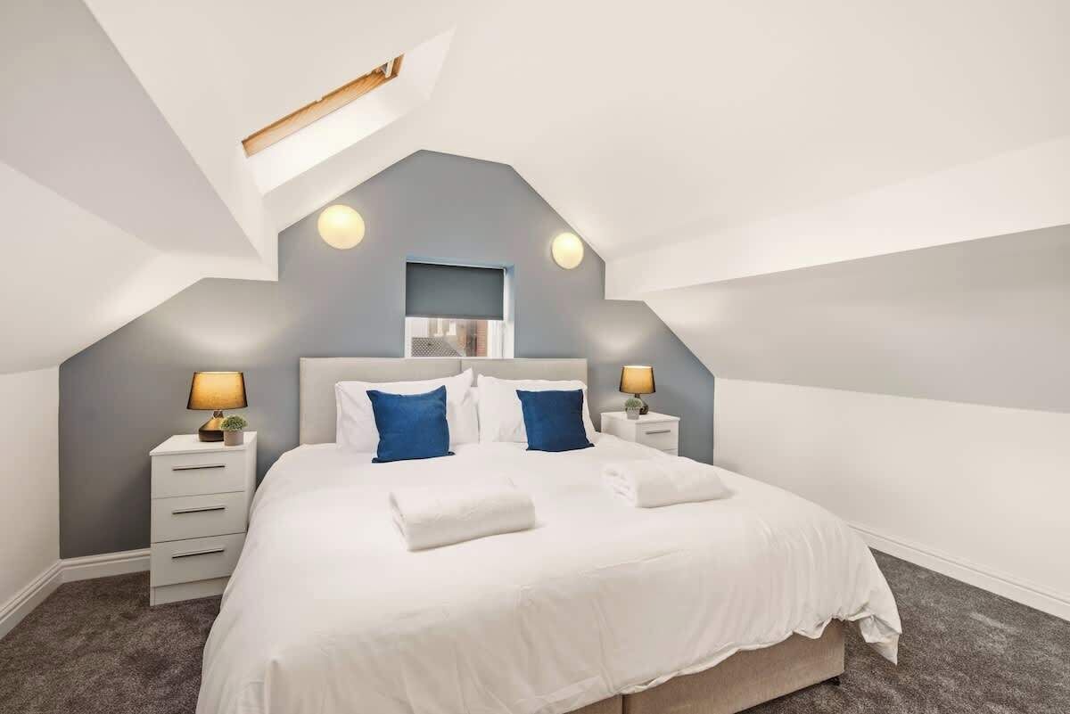 Une chambre avec un plafond en pente, un lit recouvert de draps blancs, deux tables de chevet avec des lampes et une petite fenêtre.