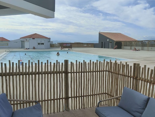Ein Schwimmbecken mit Holzlattenzaun im Vordergrund, ein überdachter Bereich mit Liegestühlen und im Hintergrund Gebäude mit roten Ziegeldächern.