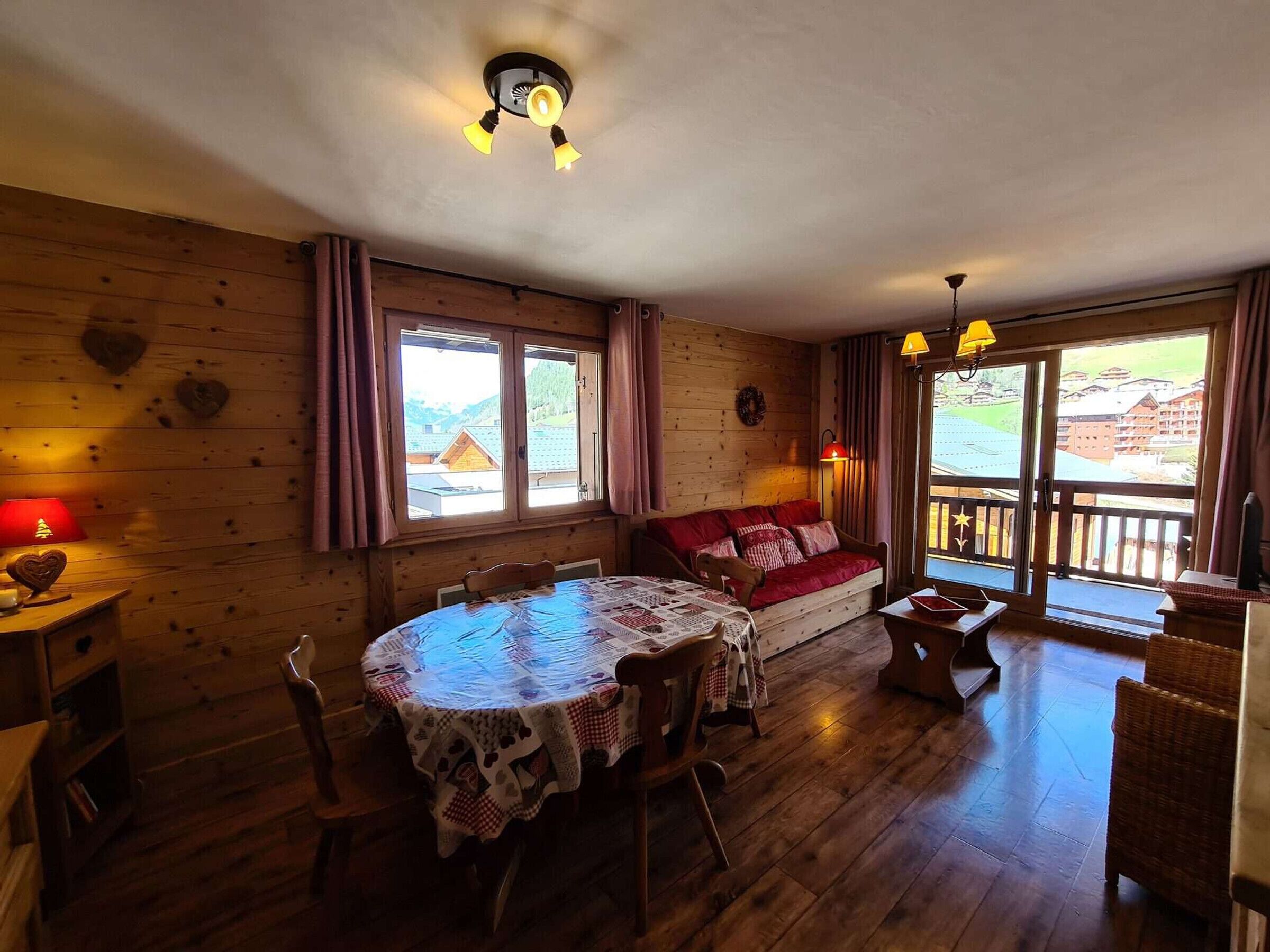Een gezellige kamer met houten wanden, een ronde tafel met tafelkleed, een rode bank en uitzicht op gebouwen door grote ramen.