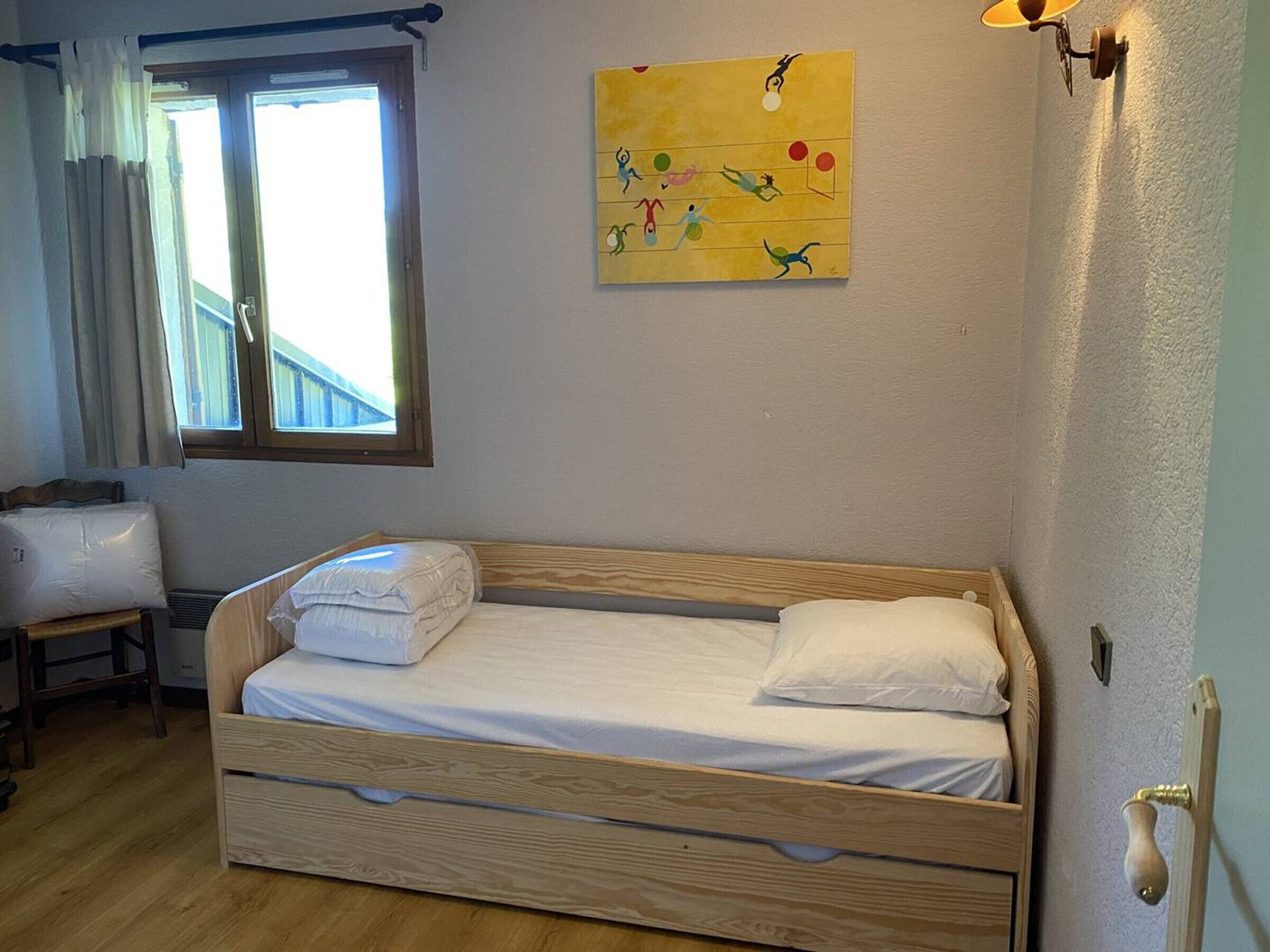 Ein Einzelbett aus Holz mit weißen Bettwäsche, ein Fenster mit Vorhängen, ein Wandbild und ein Nachttisch mit Lampe.