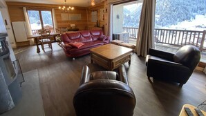 Ein gemütliches Wohnzimmer mit einem roten Ledersofa, einem Holztisch und einem schwarzen Sessel. Es gibt eine Essecke mit Holztisch und Stühlen sowie eine Küchenzeile mit Kühlschrank und Herd. Große Fenster bieten Blick auf schneebedeckte Bäume.