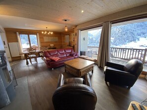 Una sala de estar acogedora con un sofá de cuero rojo, una mesa de centro de madera y un sillón negro. Hay un comedor con una mesa y sillas de madera, y una cocina con un refrigerador y estufa. Grandes ventanales ofrecen vistas a árboles cubiertos de nieve.