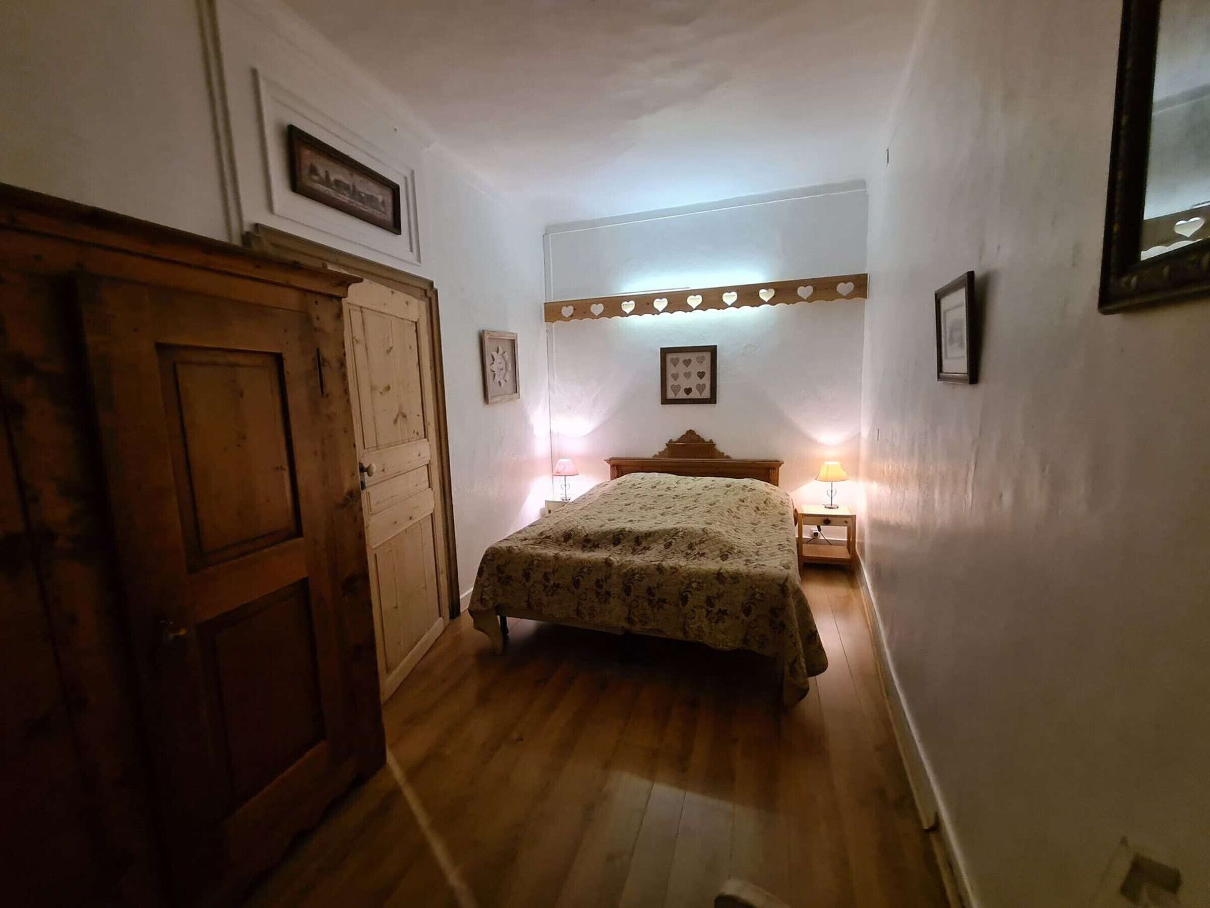 A bedroom with a wooden door, a bed with a floral bedspread, two bedside lamps, and a wall-mounted light fixture.
