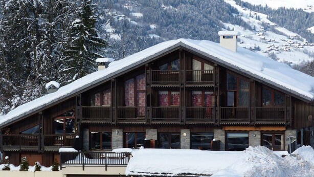 En chalet-lignende bygning med flere balkoner, en synlig butiksvindue og sneklædte bjerge i baggrunden.