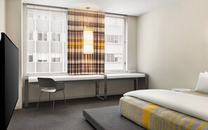Habitación de hotel con una cama, un escritorio y una silla. Cuenta con una ventana con persianas y cortina.