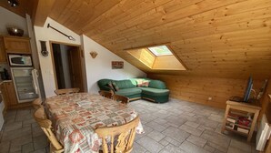 Ein gemütliches Dachzimmer mit Holzbalkendecke, einem Esstisch mit Stühlen, einem grünen Sofa und einer kleinen TV-Konsole.