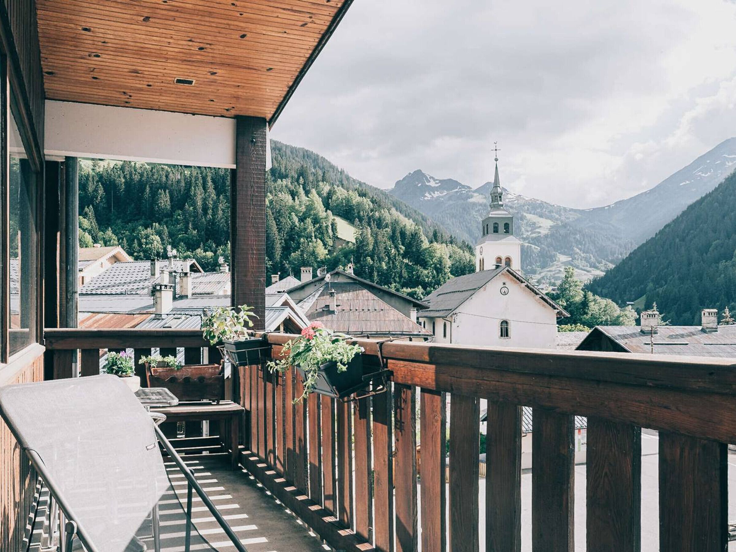 A balcony with a wooden railing overlooking a mountainous landscape with a church and residential buildings.