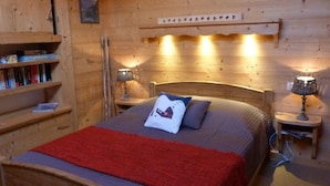 Ein Zimmer mit Holzboden, einem Bett, zwei Nachttischen mit Lampen, einem Bücherregal und einer Wandablage.