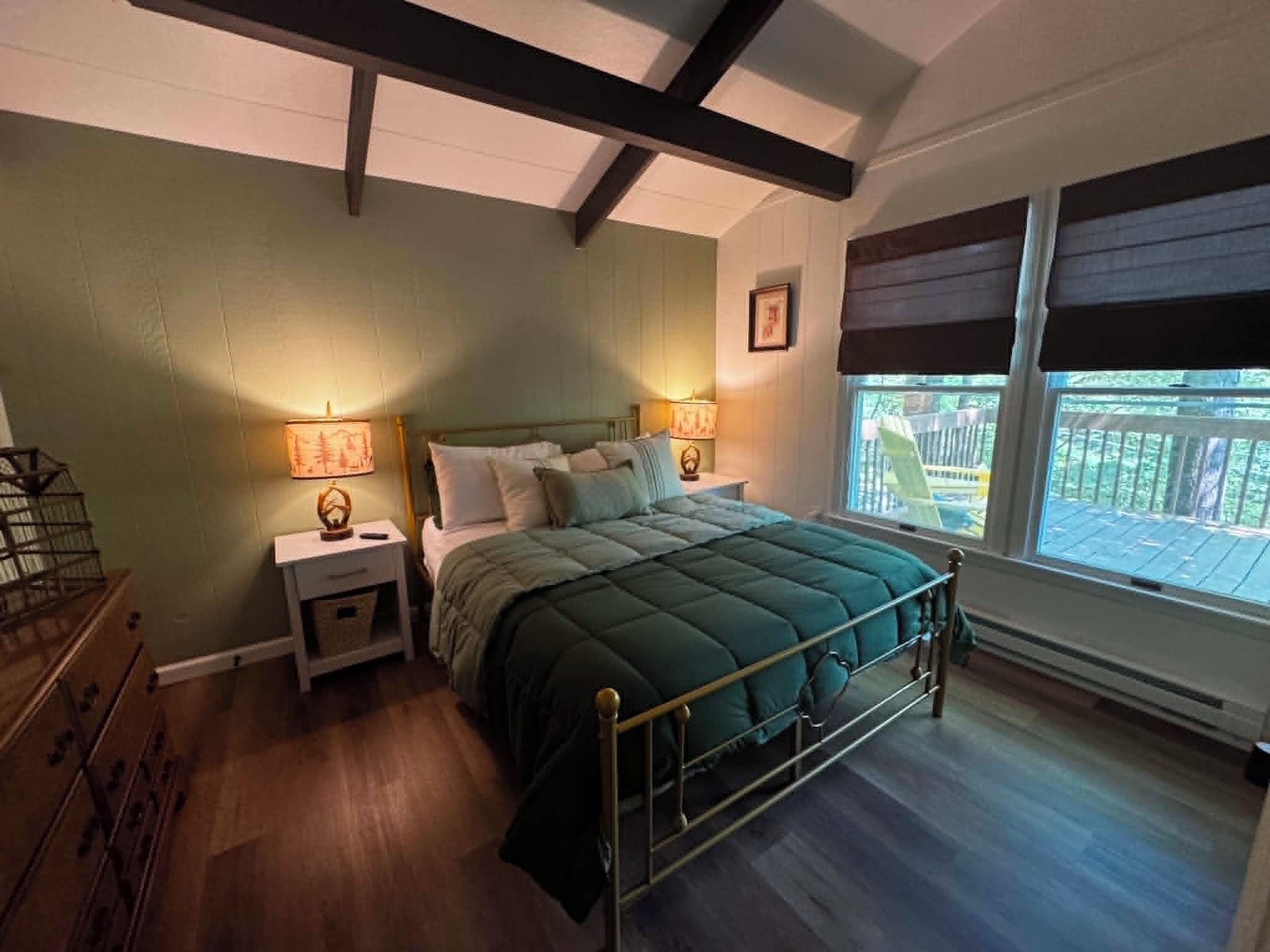 Un dormitorio con estructura de cama metálica, dos mesitas de noche con lámparas, una ventana con vistas al exterior y suelo de madera.