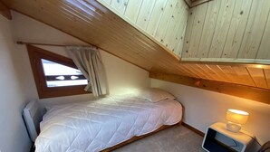 Um quarto pequeno e acolhedor com uma cama de solteiro, uma janela com cortinas, uma mesa de cabeceira com abajur e um radiador.