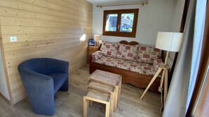 Een kamer met een houten wand, een bank met gekleurde kussens, een blauwe stoel, een klein houten tafeltje en een raam met uitzicht op gebouwen.
