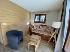 A room with a wooden wall, a sofa with patterned cushions, a blue chair, a small wooden table, and a window with a view of buildings.