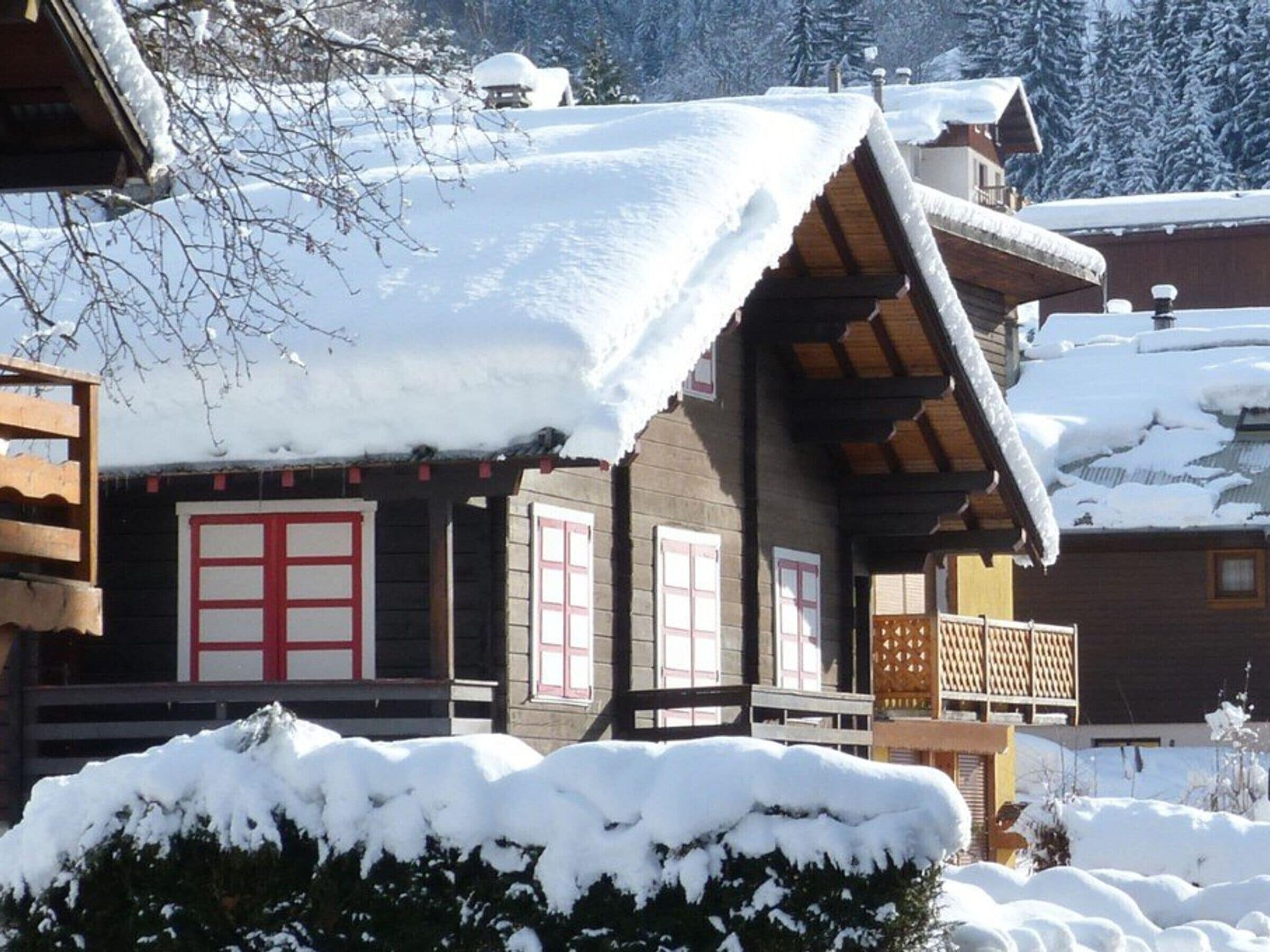 A wooden chalet with a snow-covered roof, a balcony, and a traditional red and white window design.