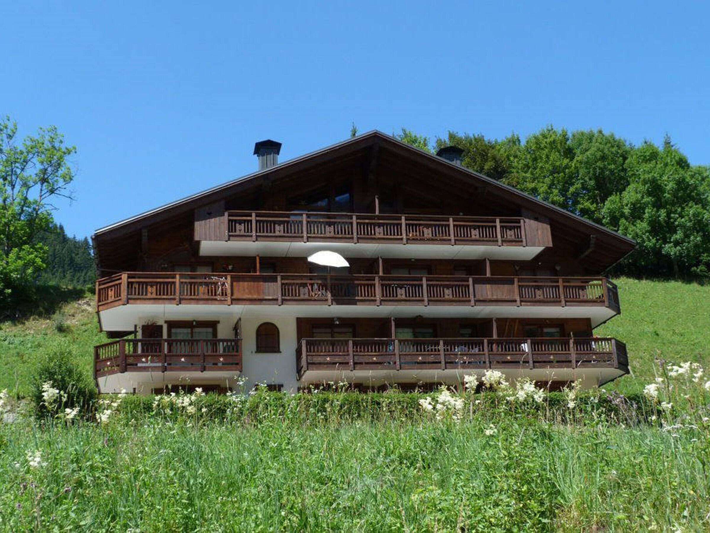 Un chalet en bois à deux étages, doté de balcons, situé dans un champ verdoyant avec des arbres en arrière-plan.