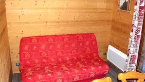 En stuga med träpanel på insidan, en röd soffa med mönster, ett träbord och ett fönster med mönstrade gardiner.