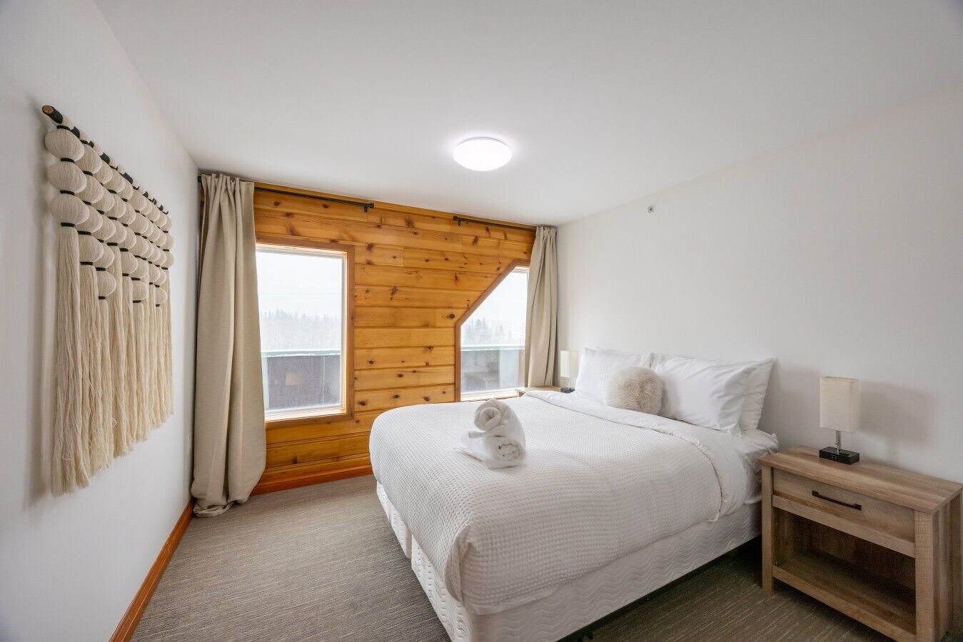 A bedroom with a large bed, a wooden ceiling, a window, and a wall-mounted macrame piece.