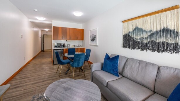 A modern living room with a grey sofa, blue chairs, a round coffee table, and a kitchen in the background. - Suite 8 at The Gem | Scenic Canmore Getaway (Harvie Heights)
