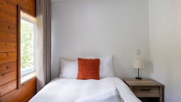 A bedroom with a wooden wall, a window, a bed with white bedding and an orange accent pillow, a bedside table with a lamp, and a nightstand with a drawer.